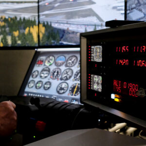 Cockpit eines Flugsimulators mit Instrumententafel, Steuerknüppel und digitaler Anzeige, im Hintergrund Flughafen und Landebahn sichtbar.