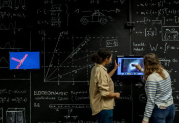 Zwei Personen vor einer Tafel mit mathematischen Formeln und Diagrammen, eine zeigt auf einen Bildschirm mit einer technischen Zeichnung.