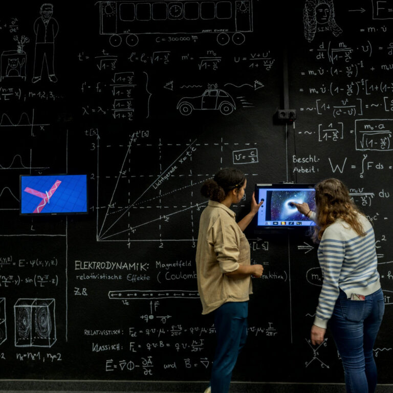 Zwei Personen vor einer Tafel mit mathematischen Formeln und Diagrammen, eine zeigt auf einen Bildschirm mit einer technischen Zeichnung.