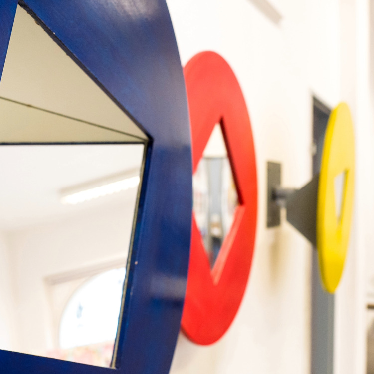 Drei runde Spiegel mit quadratischer Aussparung in den Farben Blau, Rot und Gelb an einer weißen Wand.