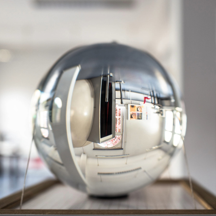 Glaskugel auf Holzsockel in einem hellen Raum mit großen Fenstern und unscharfen Personen im Hintergrund.