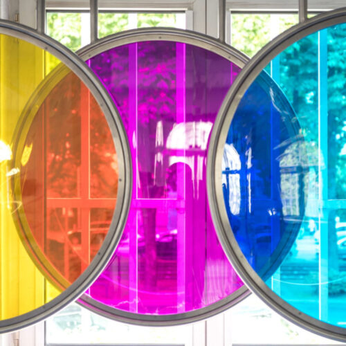 Fünf kreisrunde, durchsichtige Scheiben in Gelb, Orange, Pink, Blau und Transparent vor einem Fenster.