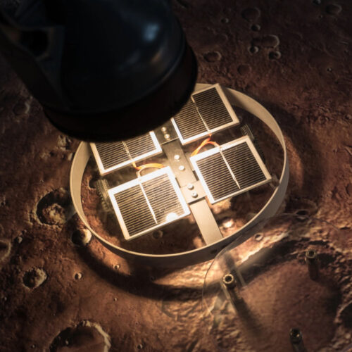 Ein rundes Solarpaneele mit quadratischen Zellen auf unebenem, kraterübersätem Boden unter einer Lampe.