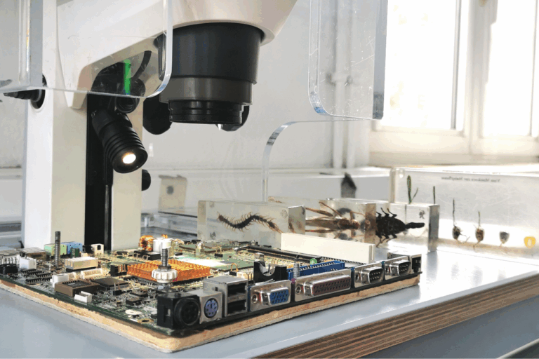 Nahaufnahme einer Platine mit verschiedenen elektronischen Bauteilen unter einer Lupe mit Beleuchtung auf einem Tisch vor einem Fenster.