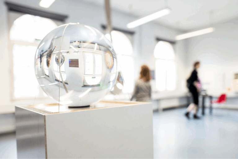 Glaskugel auf Holzsockel in einem hellen Raum mit großen Fenstern und unscharfen Personen im Hintergrund.