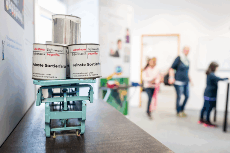 Kleiner, grüner Hubwagen mit drei weißen Dosen, darauf steht 'Abenteuer Informatik' und 'feinste Sortierfarbe', unscharfer Hintergrund mit Personen in einem hellen Raum.