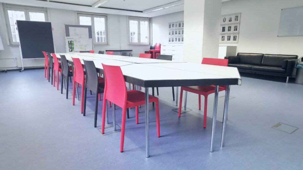 Konferenzraum mit langen Tischen, roten Stühlen, schwarzem Sofa und mehreren Fenstern.