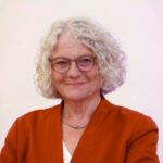 Frau mit kurzem, lockigem, weißem Haar trägt ein orangerotes Jackett und eine silberne Halskette über einem weißen Oberteil.