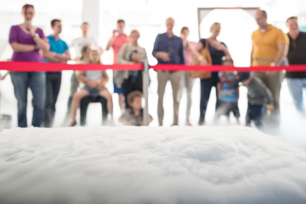 Gruppe von Menschen hinter rotem Absperrband, unscharf, im Vordergrund weiße, wolkenartige Struktur.