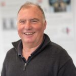 Mann mit grauem Pullover mit Reißverschluss steht vor einer unscharfen Wand mit Text und Bildern.