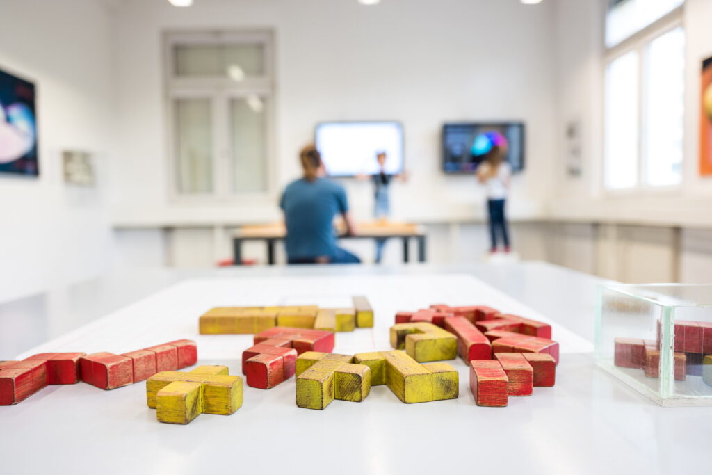 Rote und gelbe Holzbausteine in verschiedenen Formen auf weißer Oberfläche, unscharfer Hintergrund mit Stühlen und Tischen.