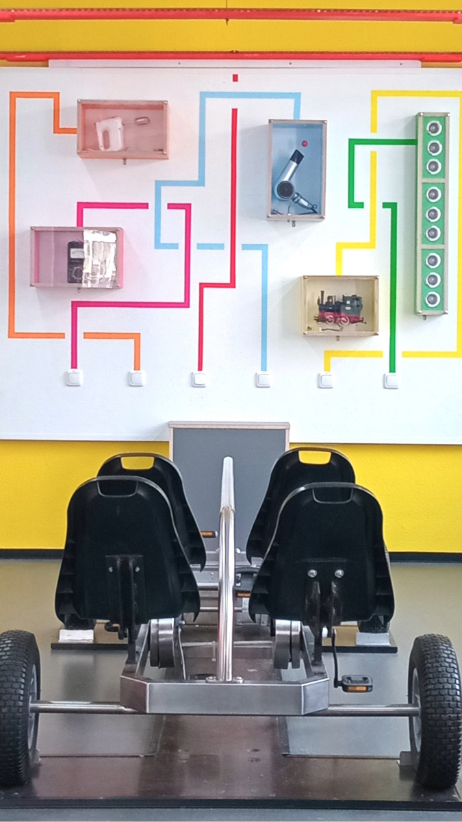 Zwei schwarze Sitze auf einem Metallgestell mit vier Reifen vor einer Wand mit farbigen Leitungen, die zu verschiedenen technischen Geräten führen.