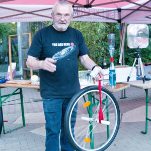 Mann steht unter einem Zelt, trägt ein schwarzes T-Shirt mit der Aufschrift 'YOU ARE HERE' und hält ein Fahrrad-Vorderrad mit roten Bändern und weißen Griffen.