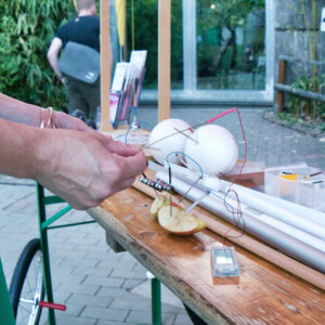 Hand mit Armbändern hält Draht, der an Apfelscheibe und kleinen elektronischen Bauteilen auf einem Holztisch befestigt ist