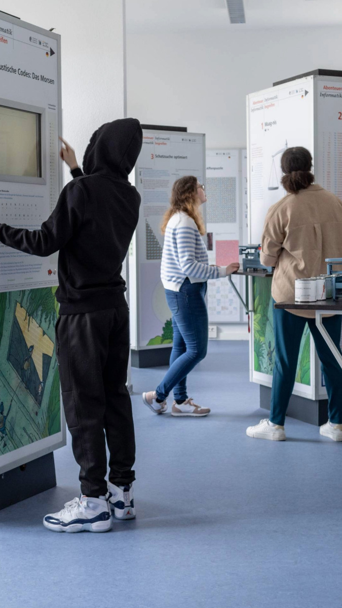 Drei Personen betrachten Ausstellungsstände mit Text und Bildern, zwei stehen, eine geht vorbei.