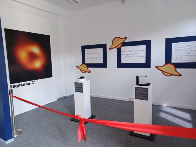 Ausstellung mit einem großen Bild des Schwarzen Lochs Sagittarius A*, drei weißen Podesten mit Beschriftungen und drei an der Wand befestigten Saturn-Symbolen, abgesperrt durch ein rotes Band.