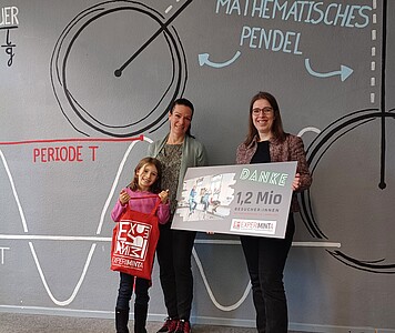 Drei Personen stehen vor einer Wand mit einer großen Zeichnung eines mathematischen Pendels und halten ein Schild mit der Aufschrift 'DANKE 1,2 Mio BESUCHER IN 2023'.