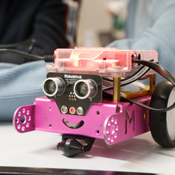 Kleiner pinkfarbener Roboter mit zwei runden Sensoren als Augen, einem lächelnden Mund und zwei schwarzen Rädern, auf einem Tisch platziert.