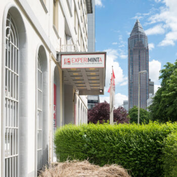 Straßenszene mit Eingang zum Experimenta Science Center, grüner Hecke und Hochhaus im Hintergrund unter blauem Himmel mit Wolken.