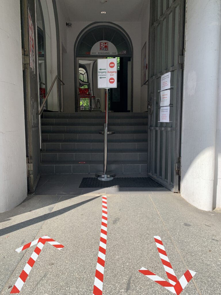 Eingang mit Treppe, darüber ein Schild mit Pfeilen und Texten 'Eingang' und 'Ausgang', auf dem Boden drei rote und weiße Pfeile, die verschiedene Richtungen anzeigen.